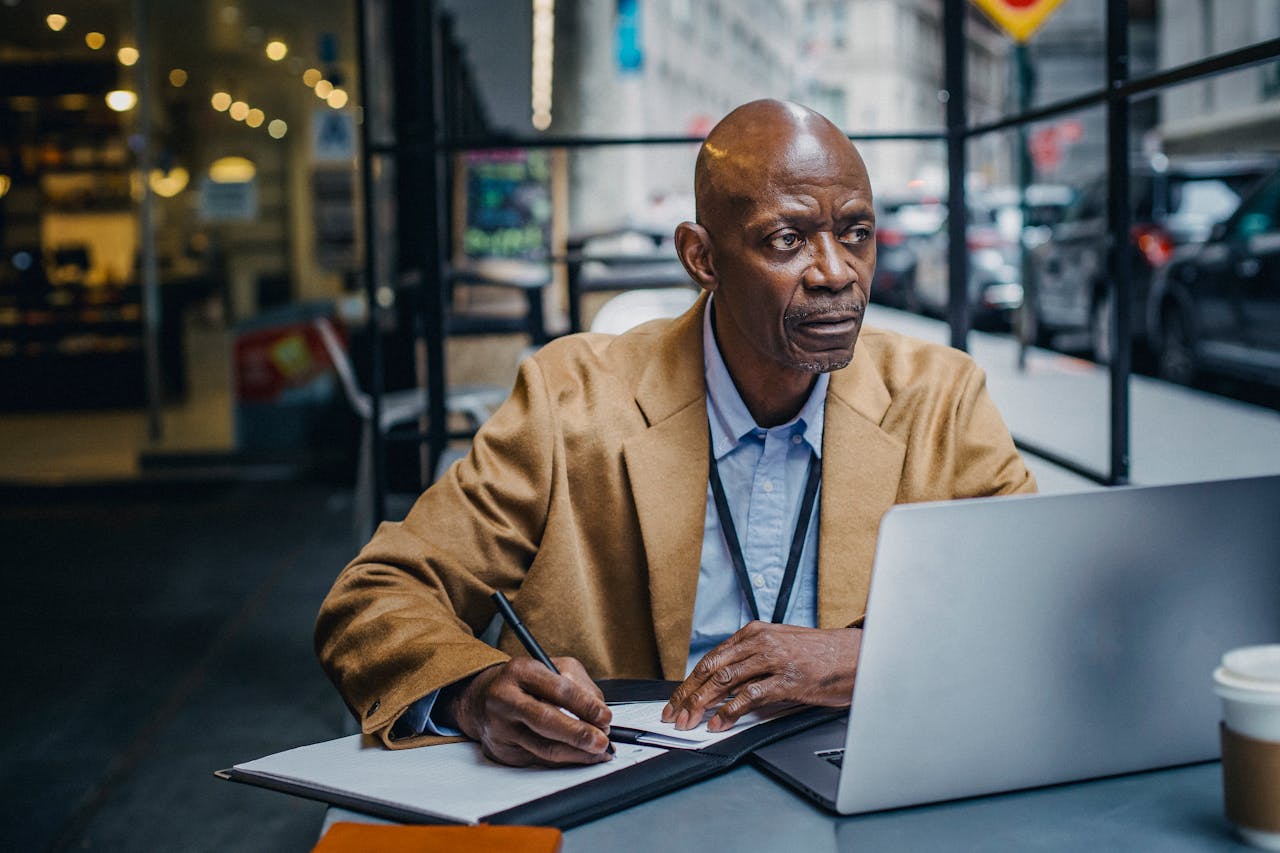 A focused man in a cafe working on his laptop and writing notes, reflecting professionalism.